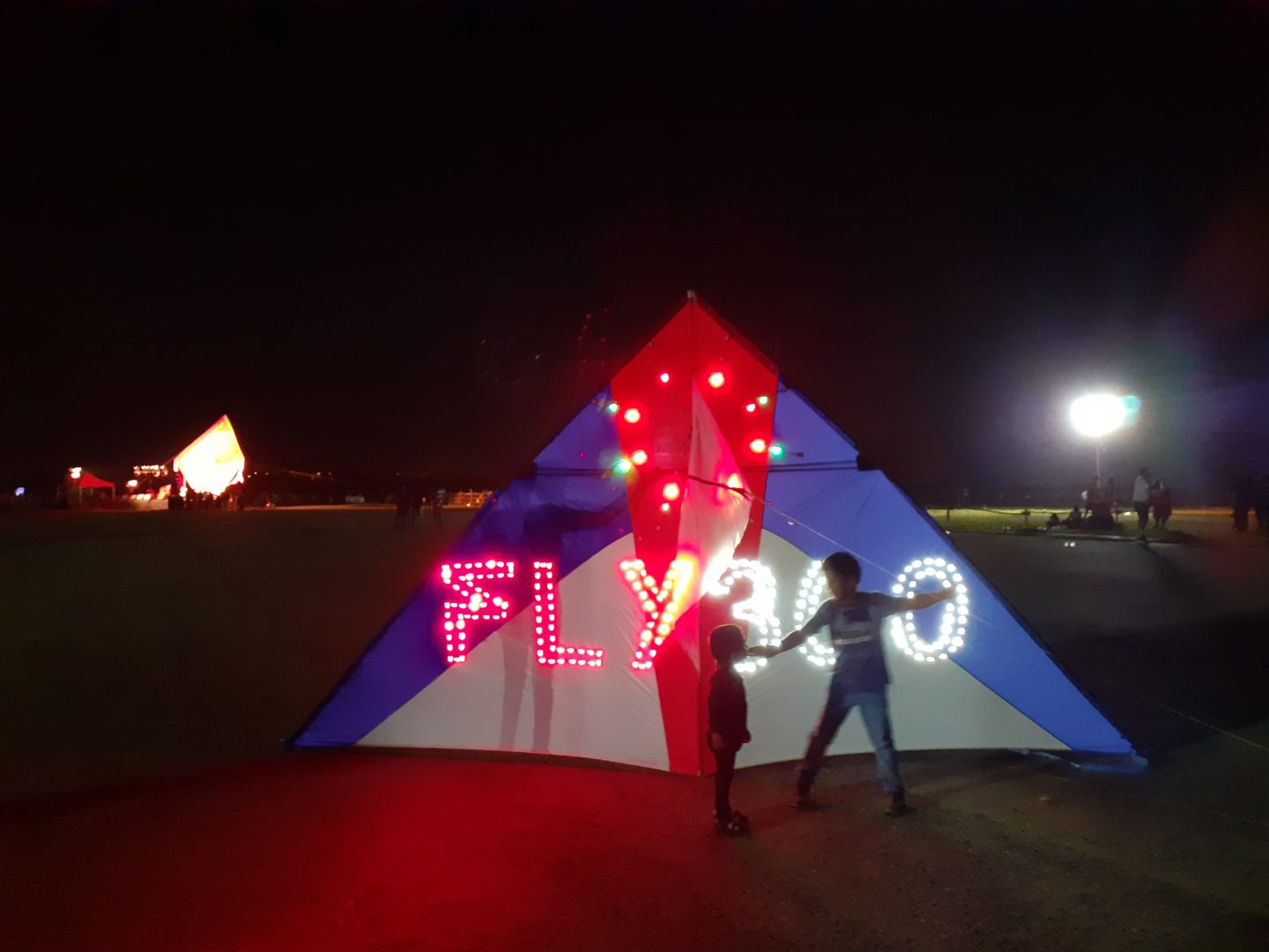 Large delta kite lit with red and white LED lights spelling FLY360, with two people standing beside it at a night kite show