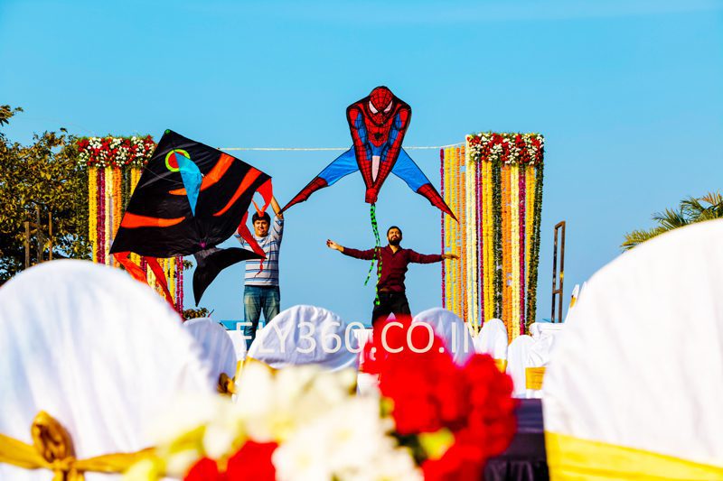 A wedding stage decorated with flowers and white drapes with colourful show kites flying above it, including a Spider-Man kite